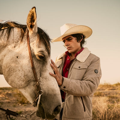 Man in a cowboy hat and coat standing next to a horse in a desert setting