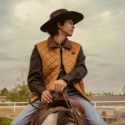 Person wearing a cowboy hat and quilted vest sitting on a horse with a cloudy sky in the background