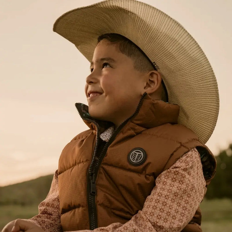 A young boy wearing a brown quilted puffer tempco vest and a large straw cowboy hat, looking up and smiling outdoors during golden hour.